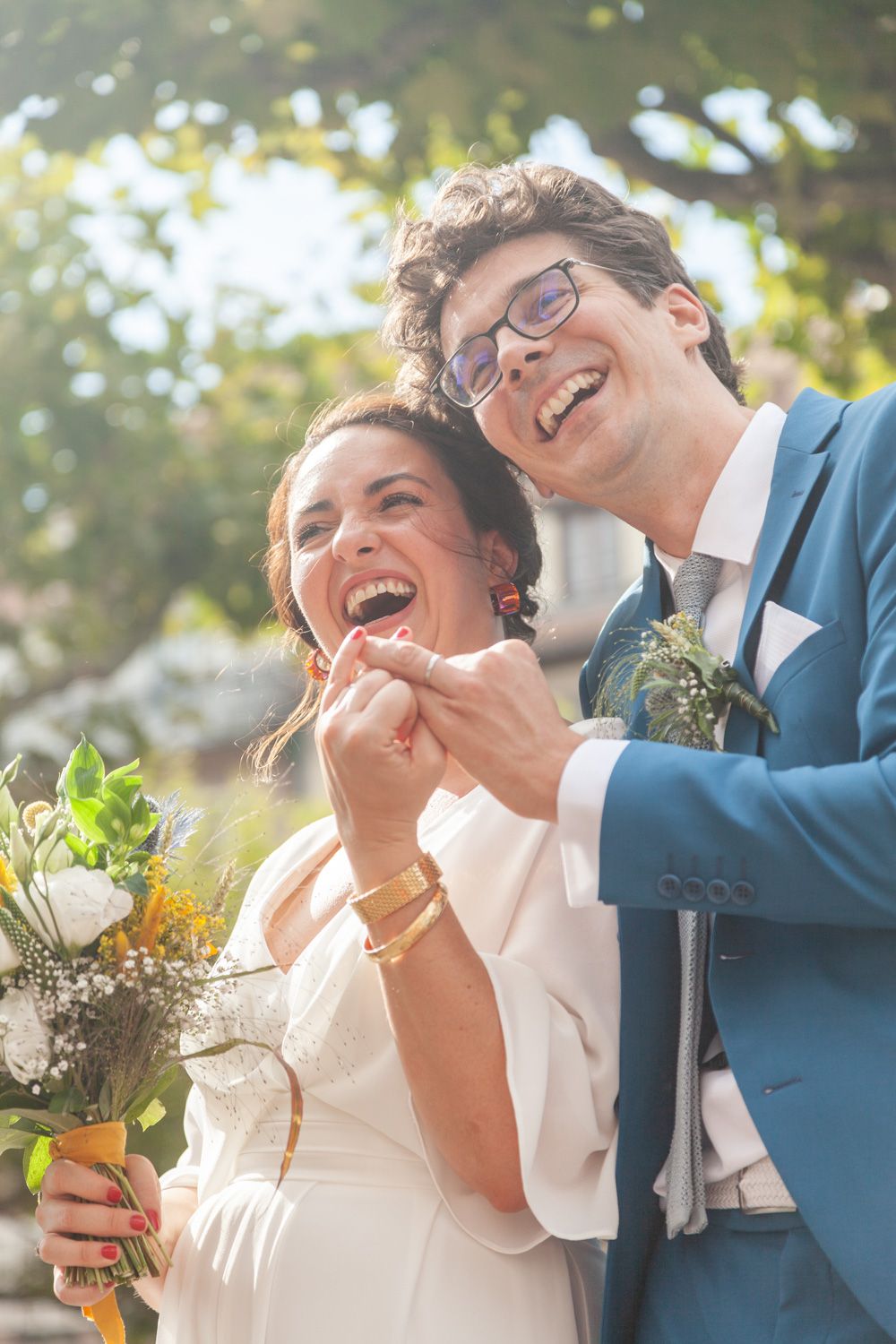 Joyful wedding couple laughing in sunlit garden setting with floral bouquet.
