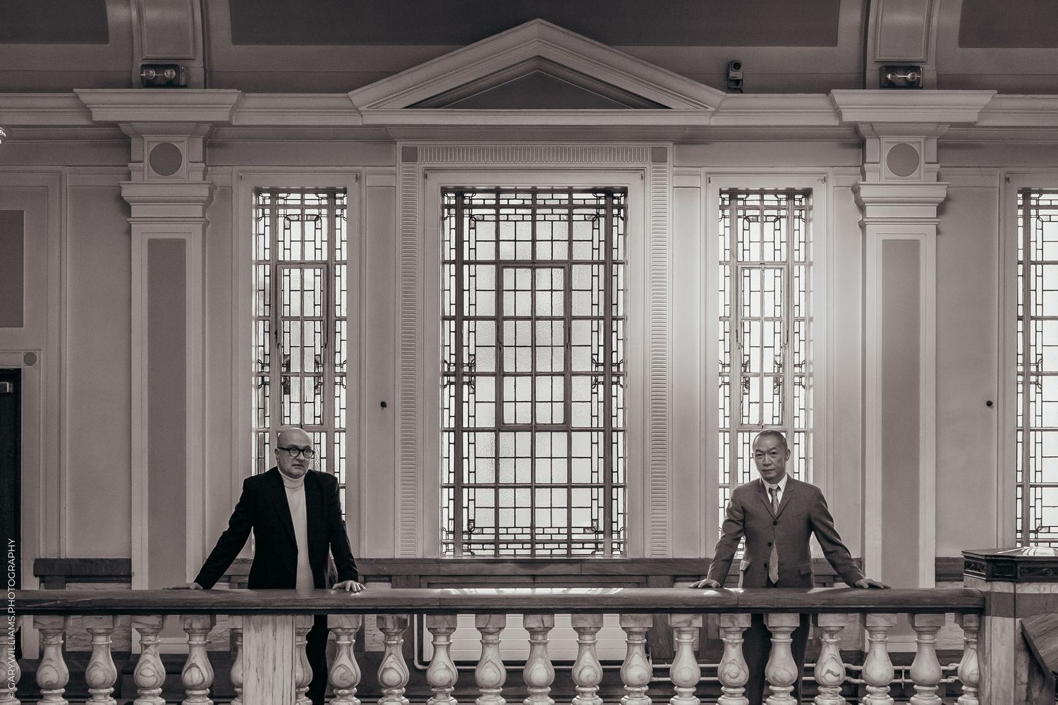 Newlywed gay couple in suits leaning on a banister in front of high windows in Islington Town Hall in London