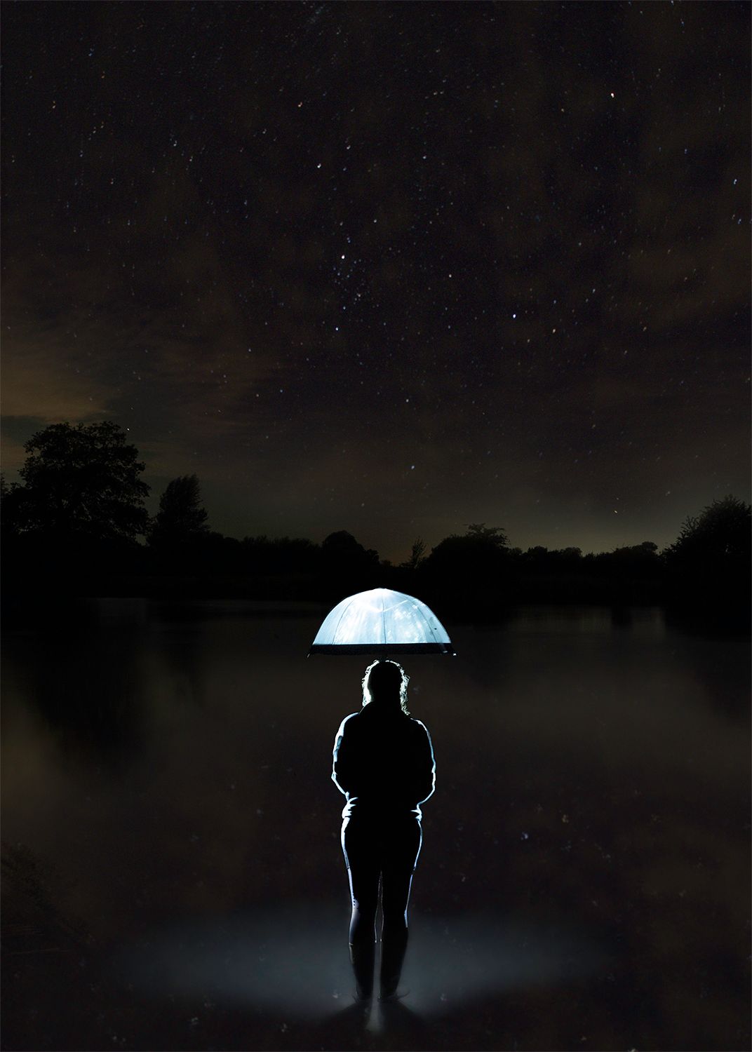 A figure outlined in light stand with a glowing umbrella in a river at night with stars shining overhead