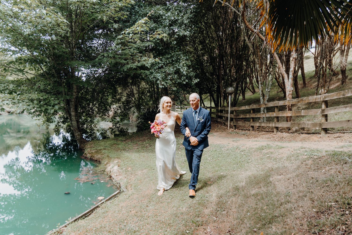 Wedding couple walks along a scenic path beside turquoise water.