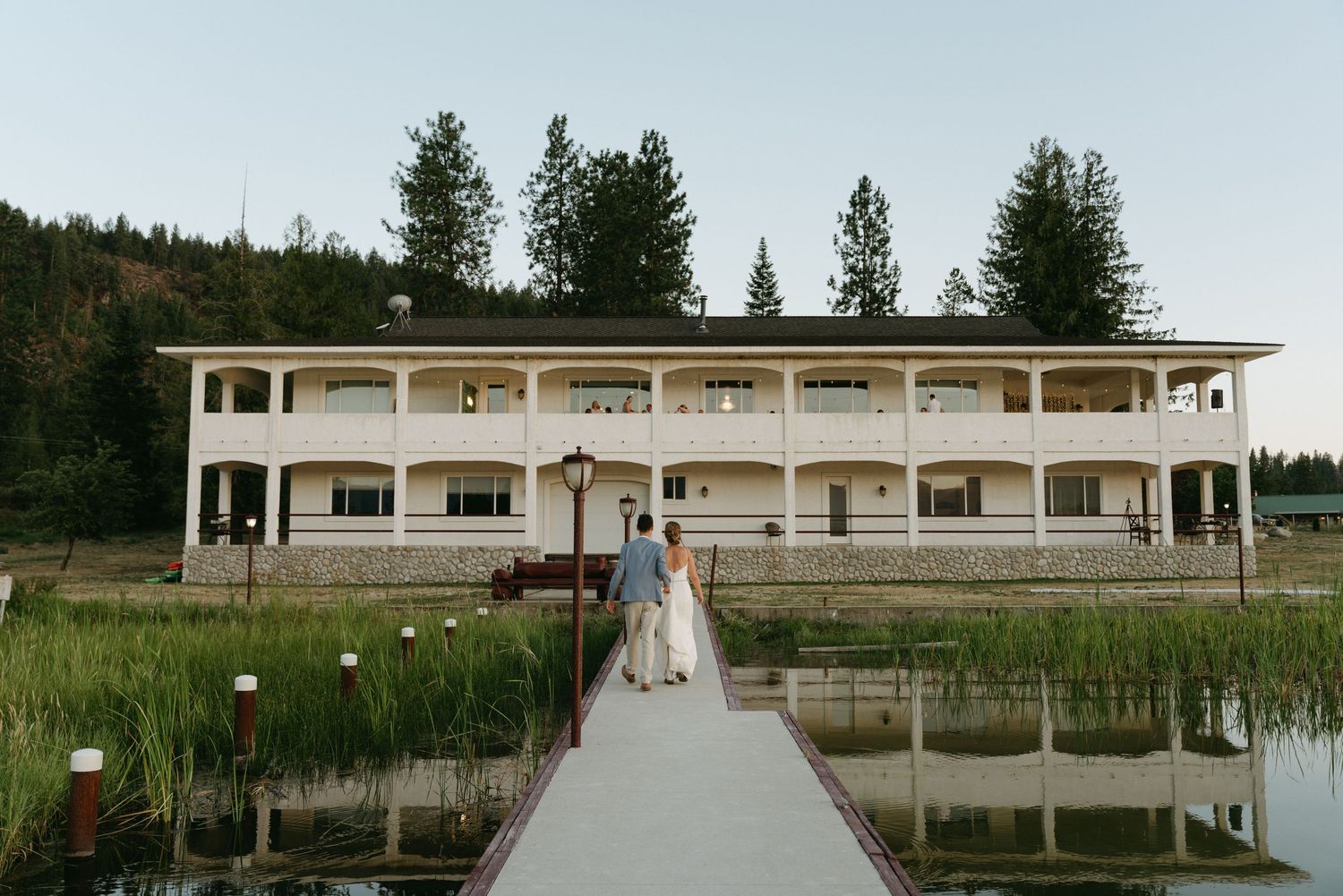 Wedding couple stands on a dock in front of a modern white venue building with pine trees in the background.