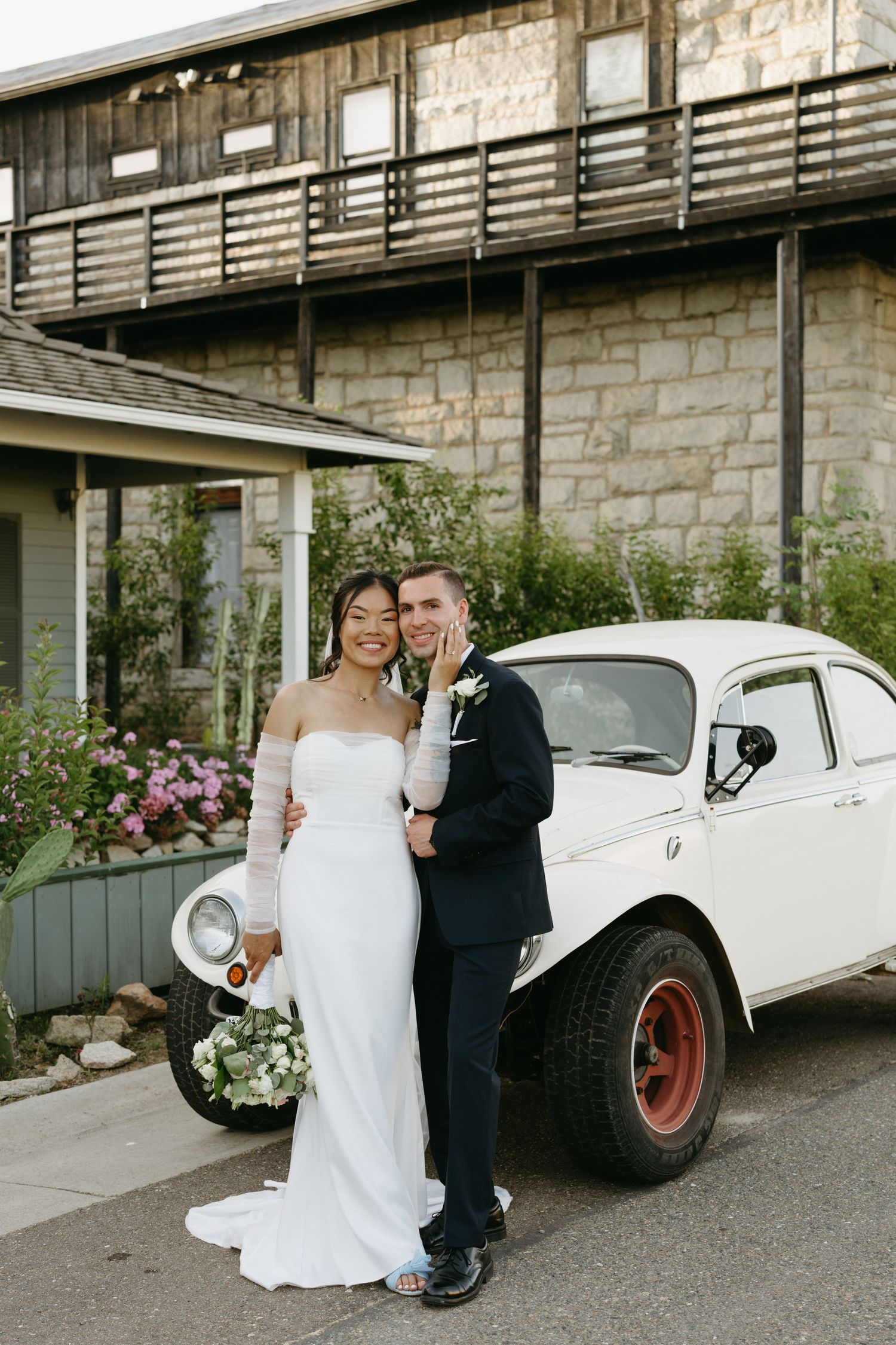 A wedding couple poses next to a vintage white Volkswagen Beetle against a rustic stone building.