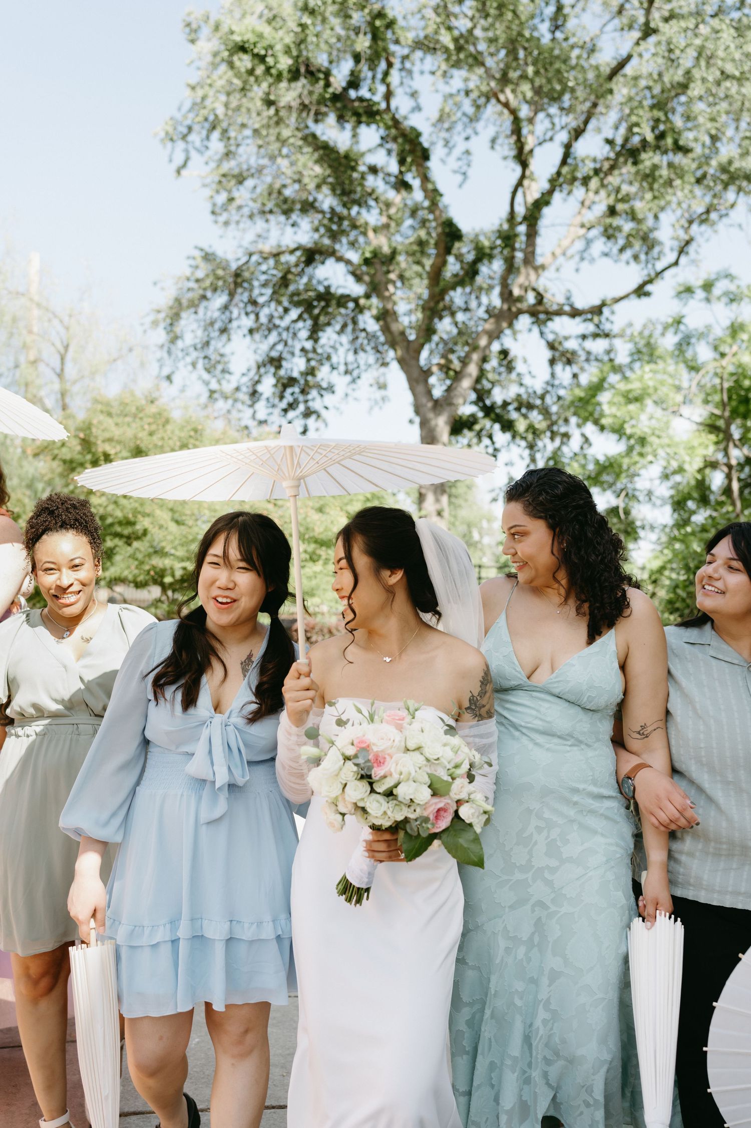 A wedding party in soft blue dresses poses together outdoors under a white parasol.