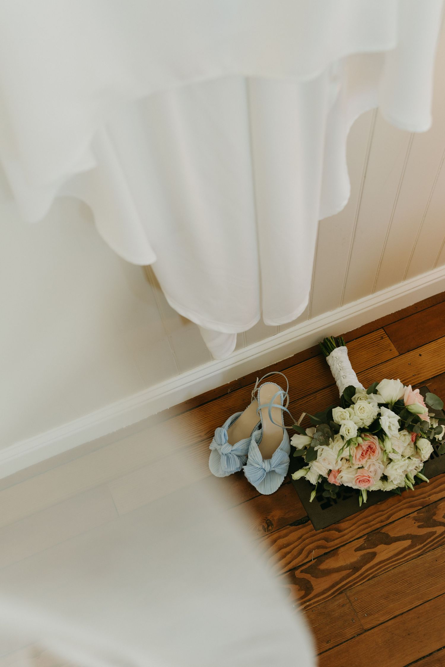 Wedding details with blue shoes and a delicate bridal bouquet resting on a wooden floor beside white fabric.