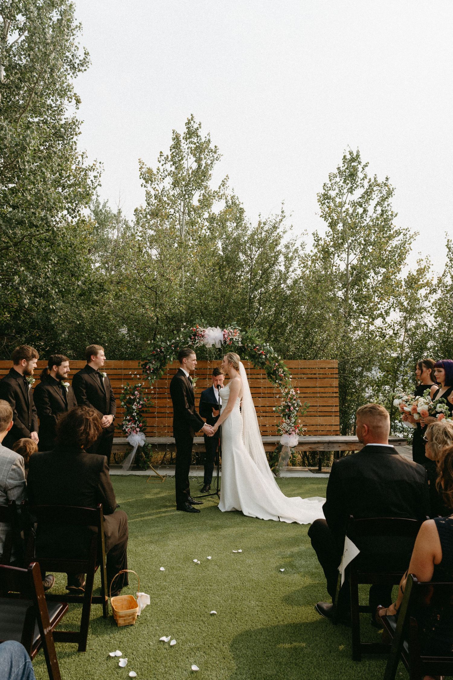 Outdoor wedding ceremony with guests seated on chairs as couple exchanges vows under wooden arch with greenery.