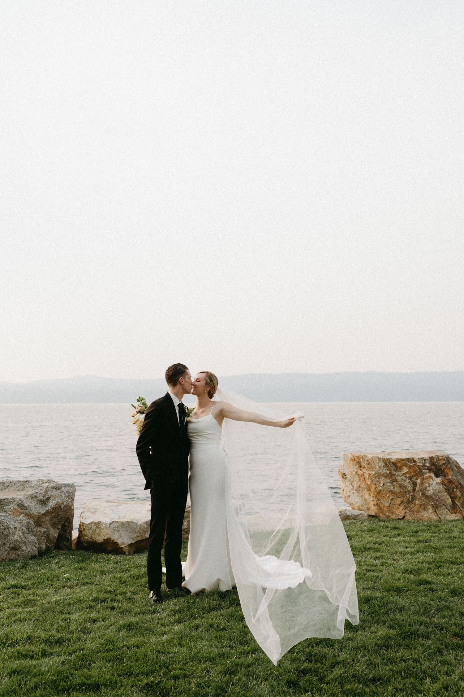 Couple in wedding attire posing with flowing veil against waterfront backdrop at sunset.