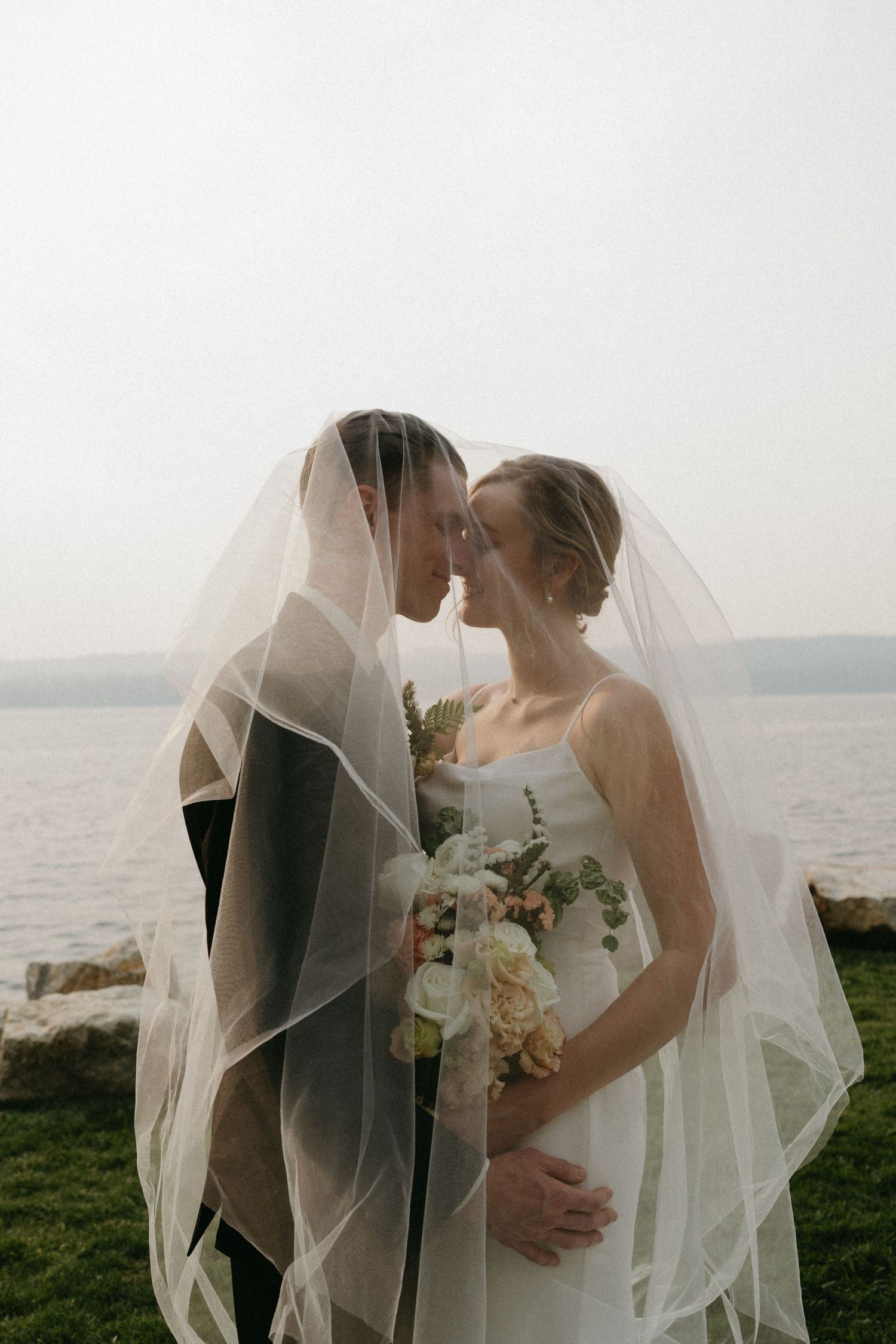 Intimate moment between newlyweds under flowing veil with waterfront view.