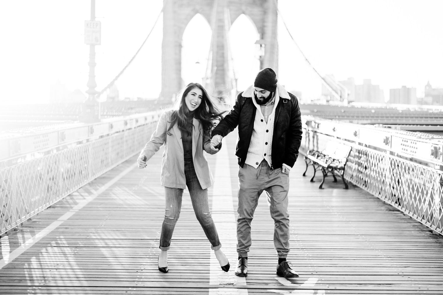 black and white image of young engaged couple laughing on an empty Brooklyn Bridge at sunrise for engagement pictures
