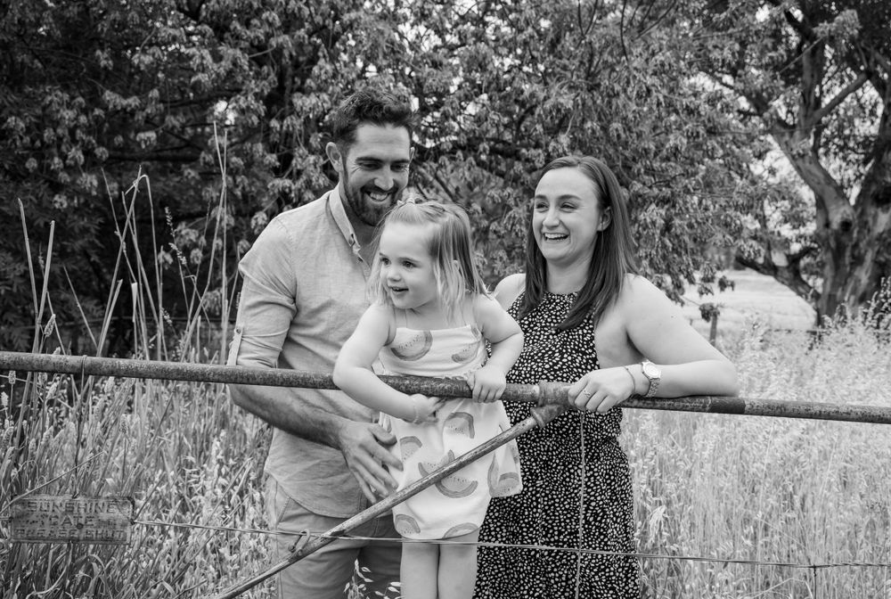 A family stands together at a wooden railing in a natural outdoor setting with tall grass and trees.