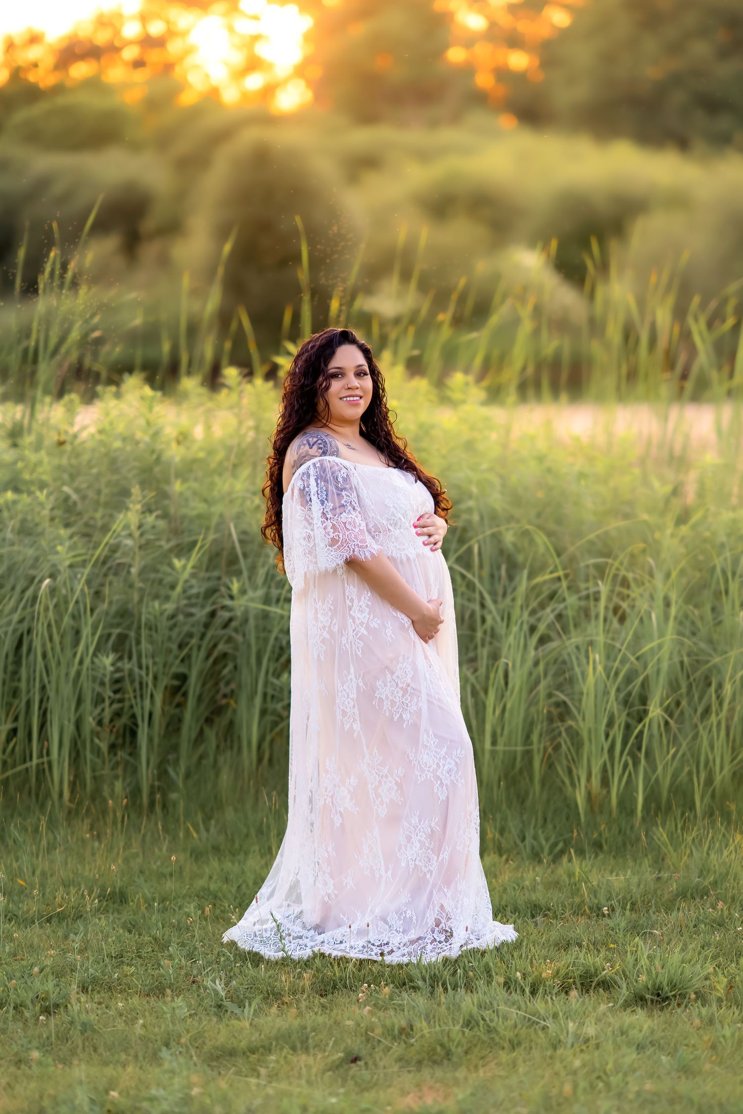 A delicate white lace maternity gown glows in golden sunset light while surrounded by tall summer grass.