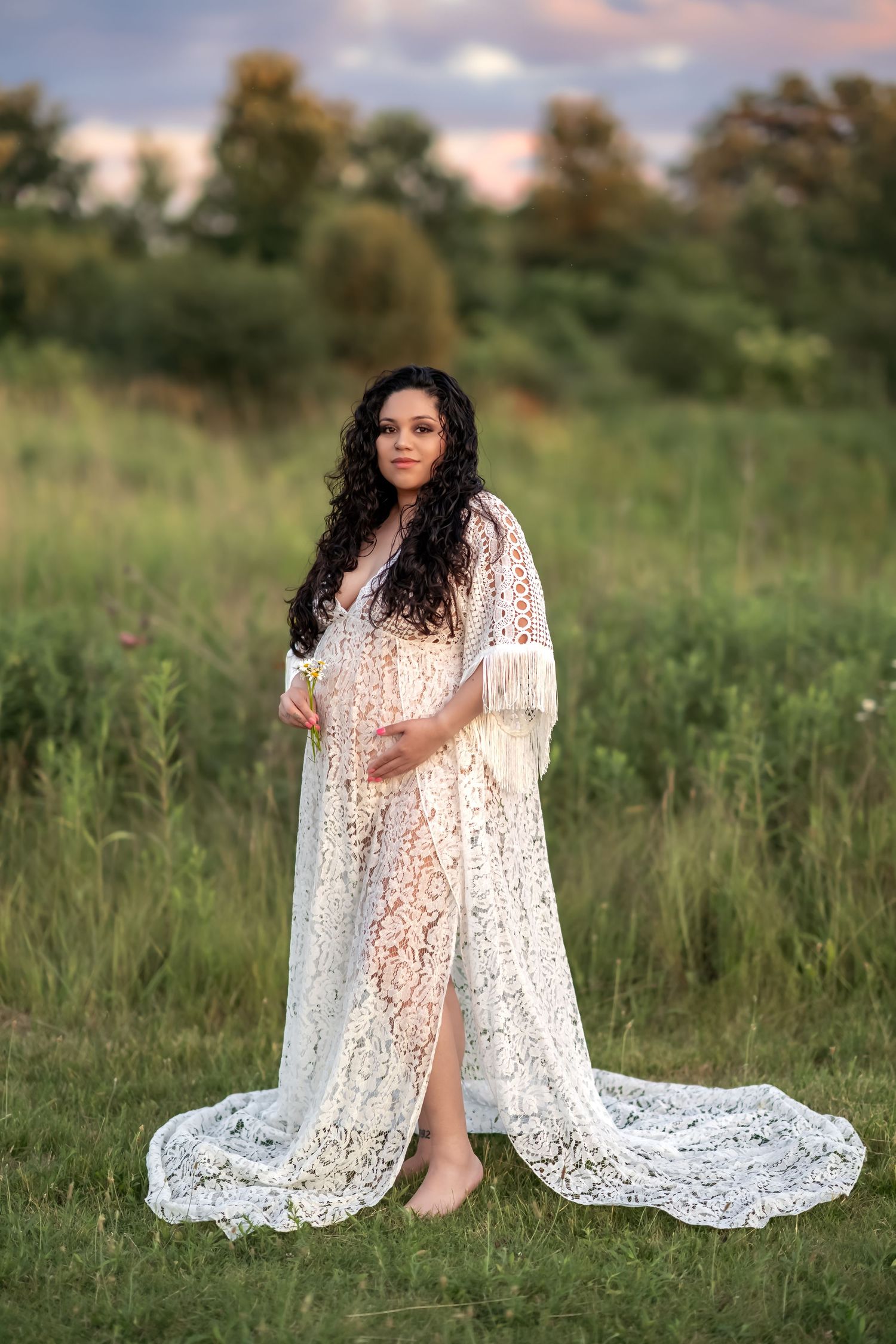 Someone in a flowing white lace dress poses gracefully in a sunlit meadow during golden hour.