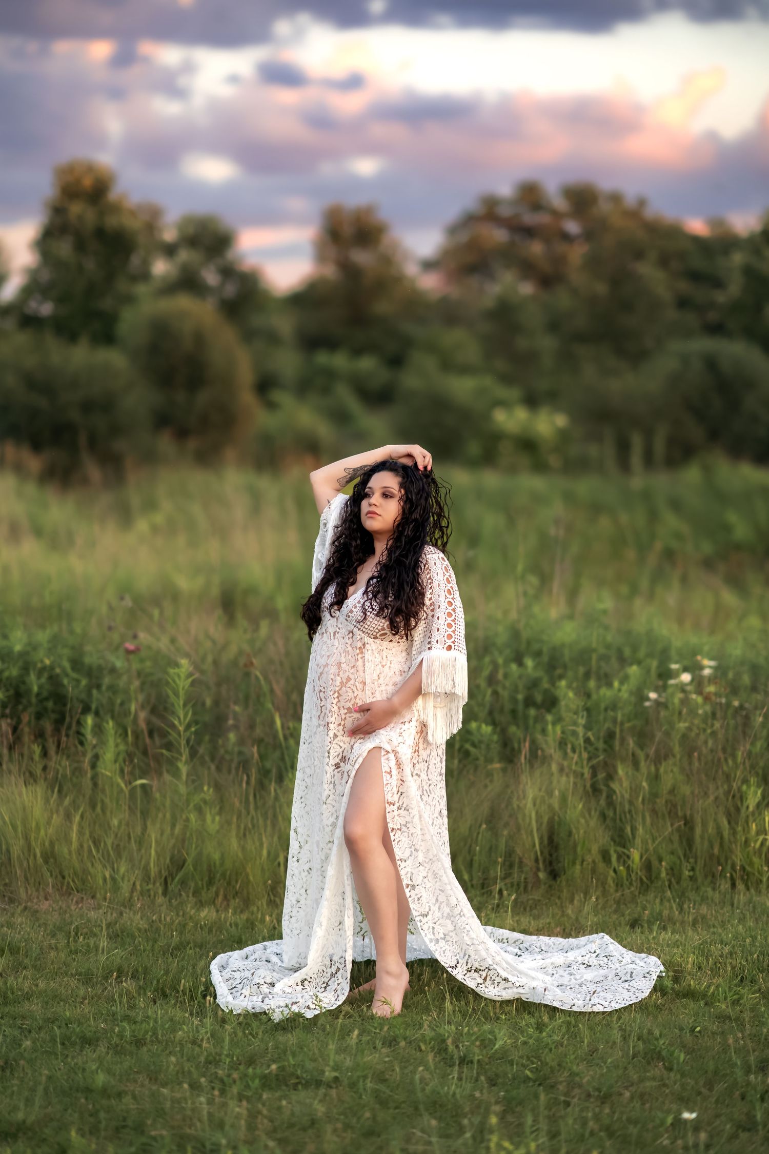 Dramatic maternity photo in white lace dress against sunset sky and tall grass field.