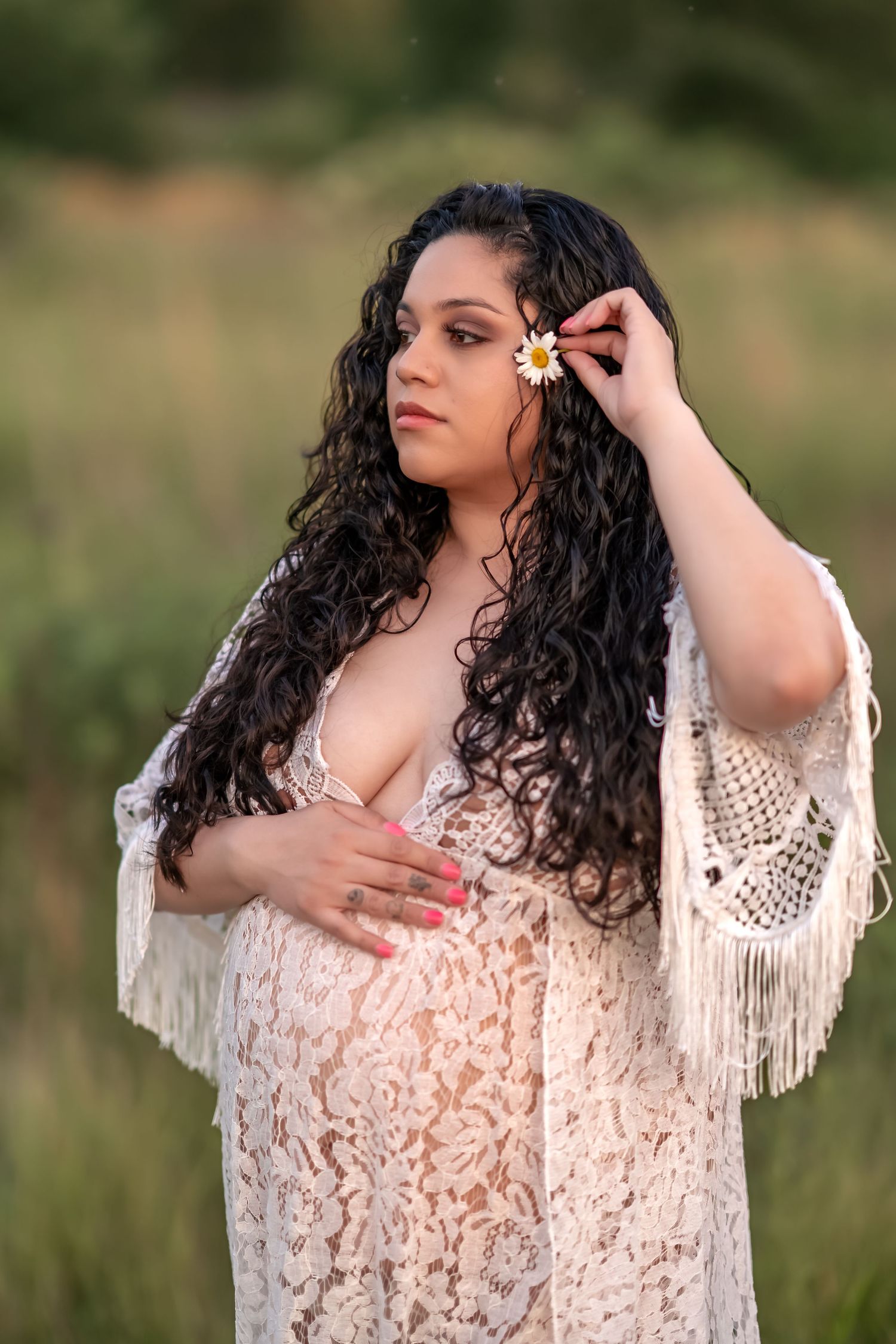 Portrait of a person with long dark curly hair wearing a cream lace dress with fringe details in an outdoor setting at sunset.