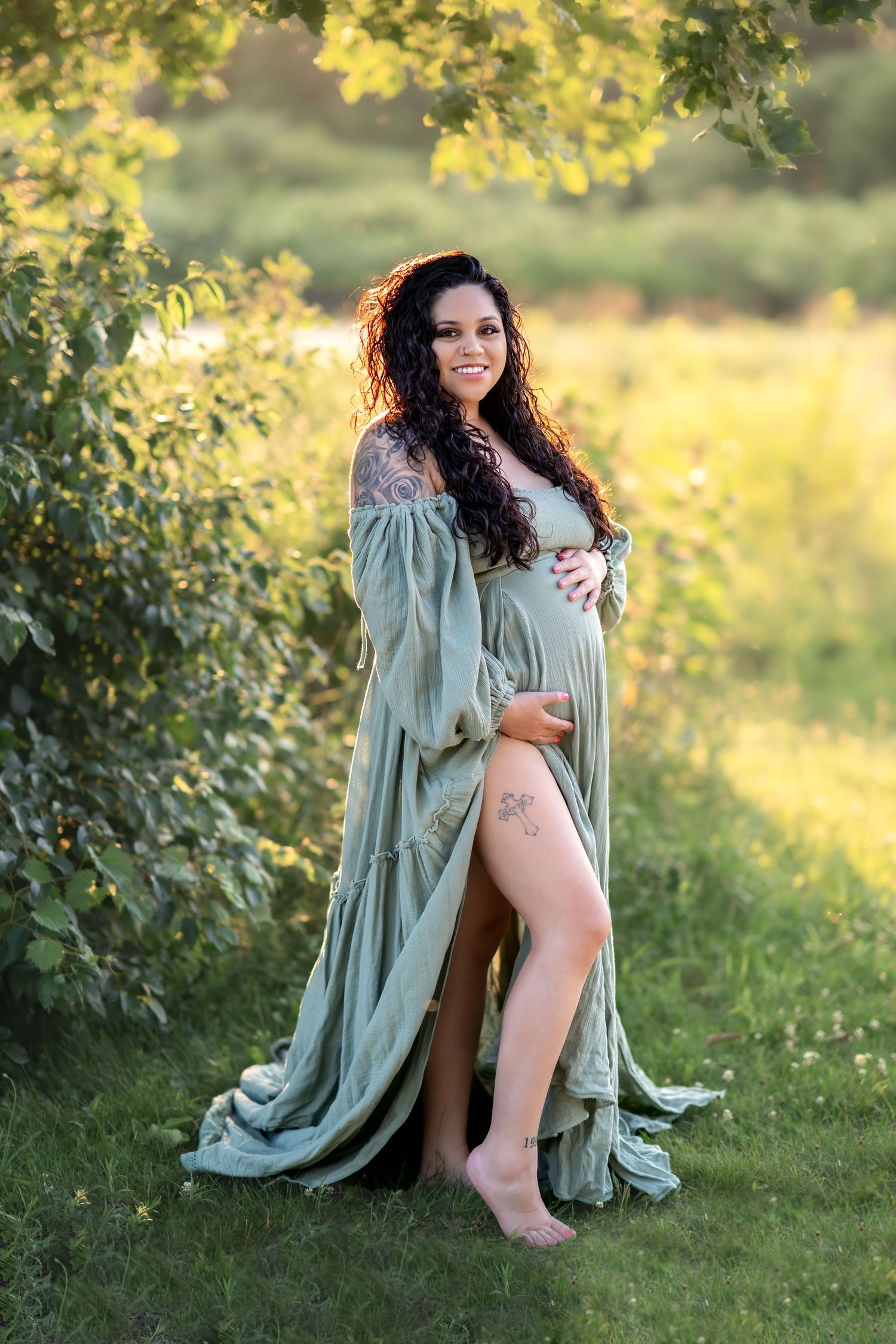 A pregnant woman in a flowing sage green dress poses outdoors in warm golden sunlight under tree branches.