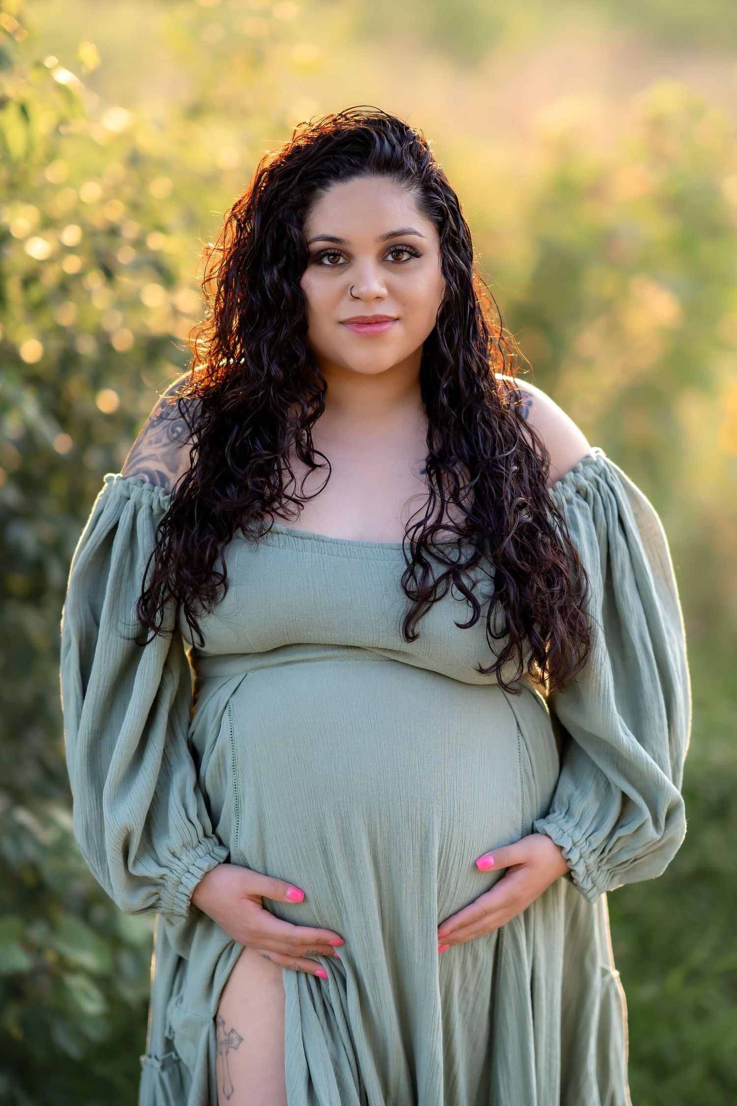 Maternity portrait in a sage green off-shoulder dress standing in golden sunlit garden setting.