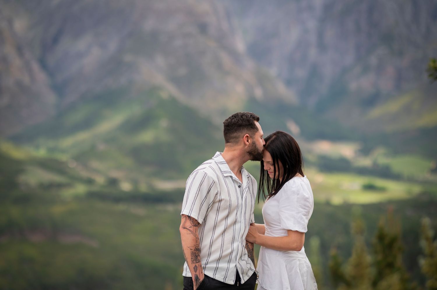 A couple shares an intimate moment against the backdrop of majestic green mountains and valleys.