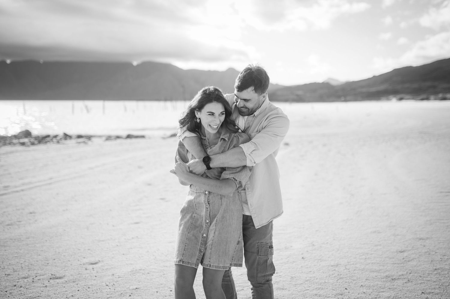 A couple embraces on a beach with mountains in the background in black and white photography.