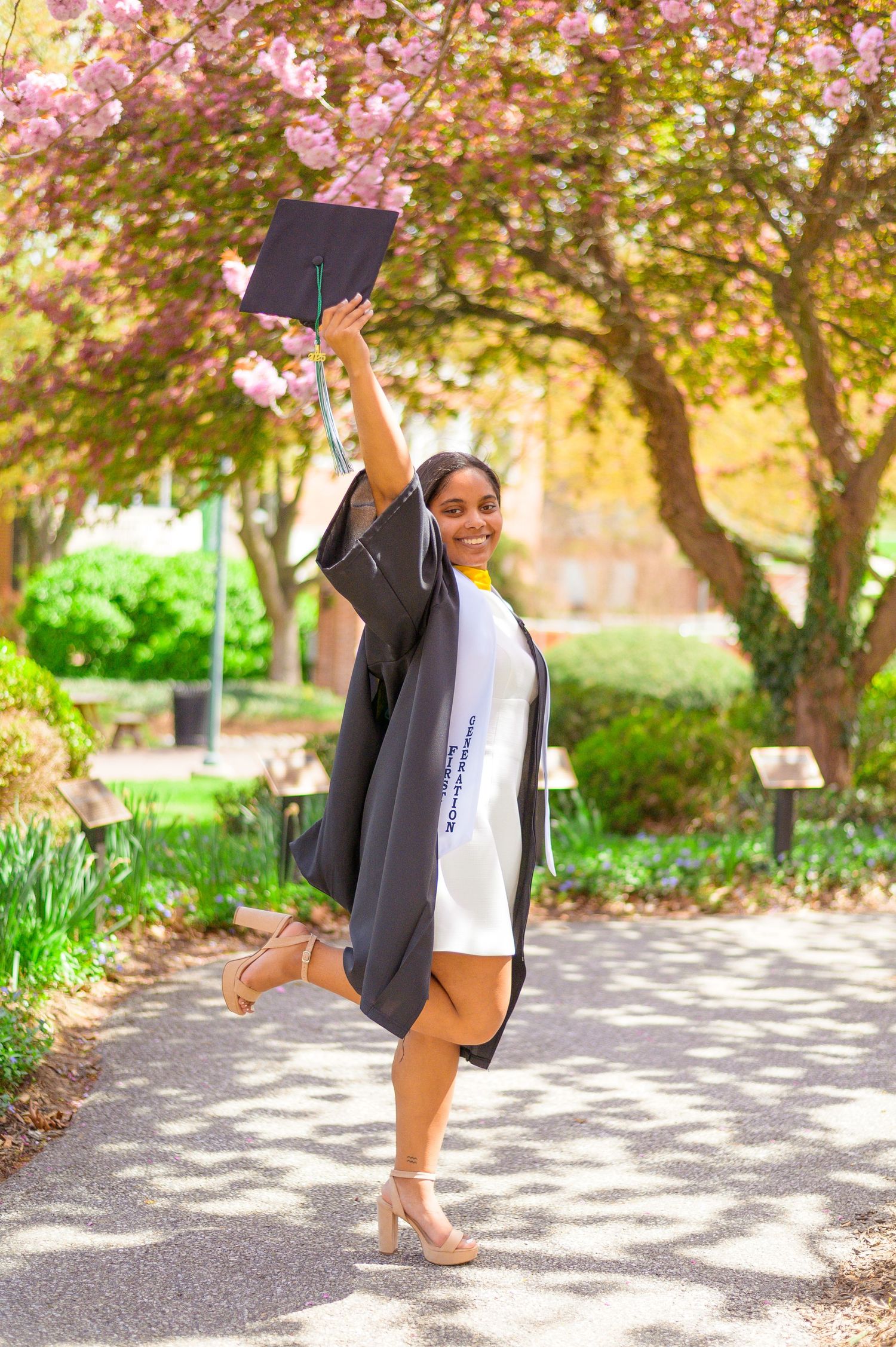 Graduate in black cap and gown celebrating with raised diploma under cherry blossoms.