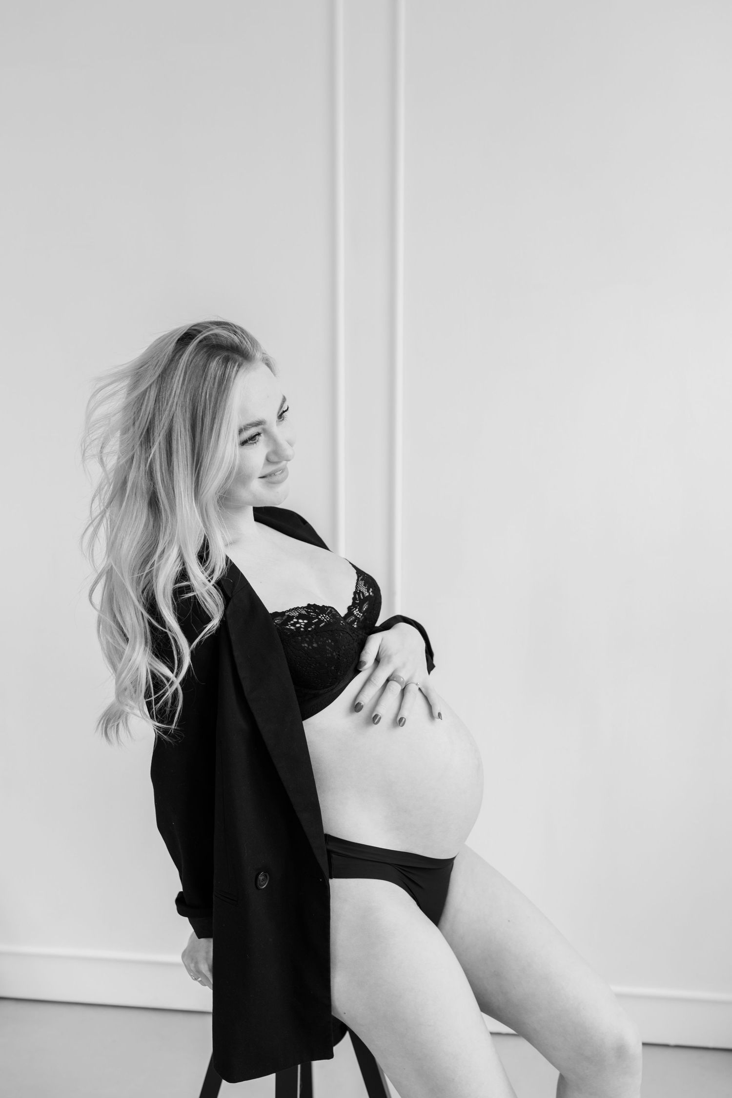 Black and white artistic boudoir photo showing figure in lingerie and open blazer against white wall.