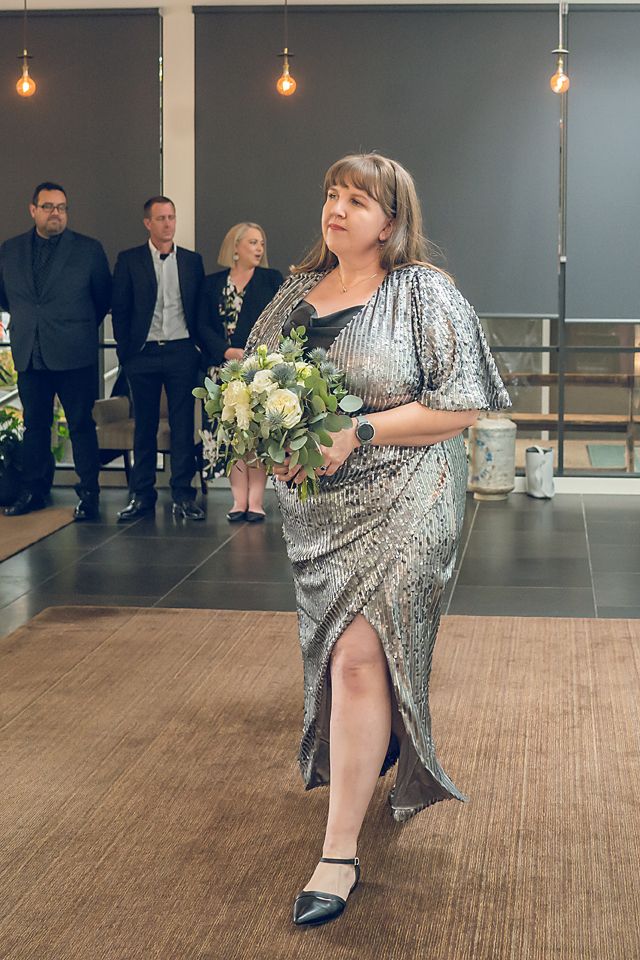 Attendee in sparkly silver dress carries white floral bouquet while walking on wooden floor at formal event.