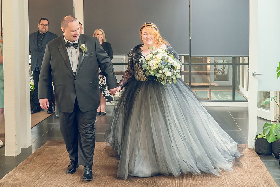 A couple in formal attire stands together at what appears to be a wedding, with the bride wearing a gray tulle gown.