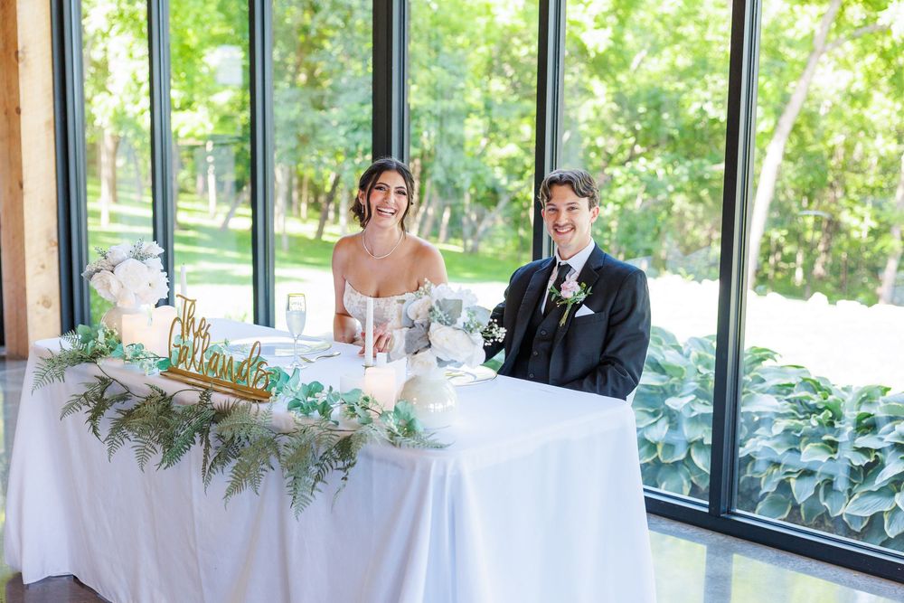 Wedding party seated at a long white decorated reception table with lush greenery arrangements.