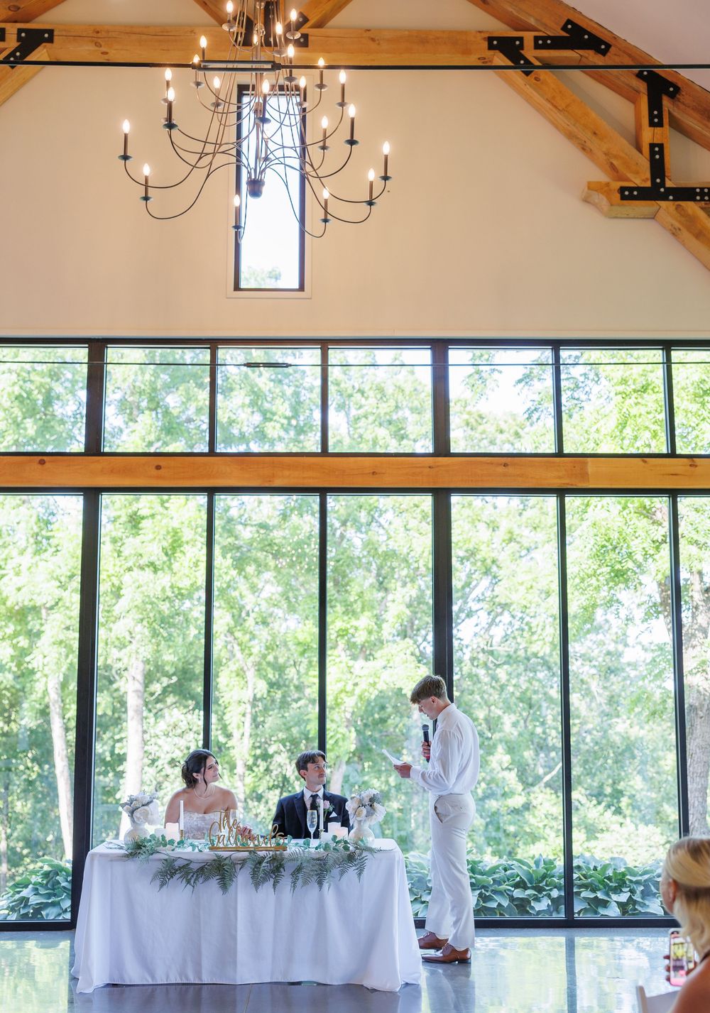 Wedding reception venue features exposed wooden beams, chandeliers, and large windows overlooking lush greenery.