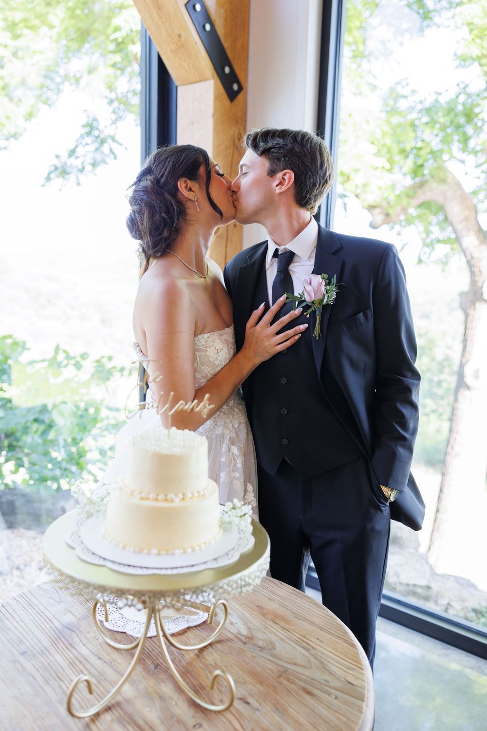 Romantic bridal couple shares intimate kiss next to elegant white wedding cake with decorative base and window view.