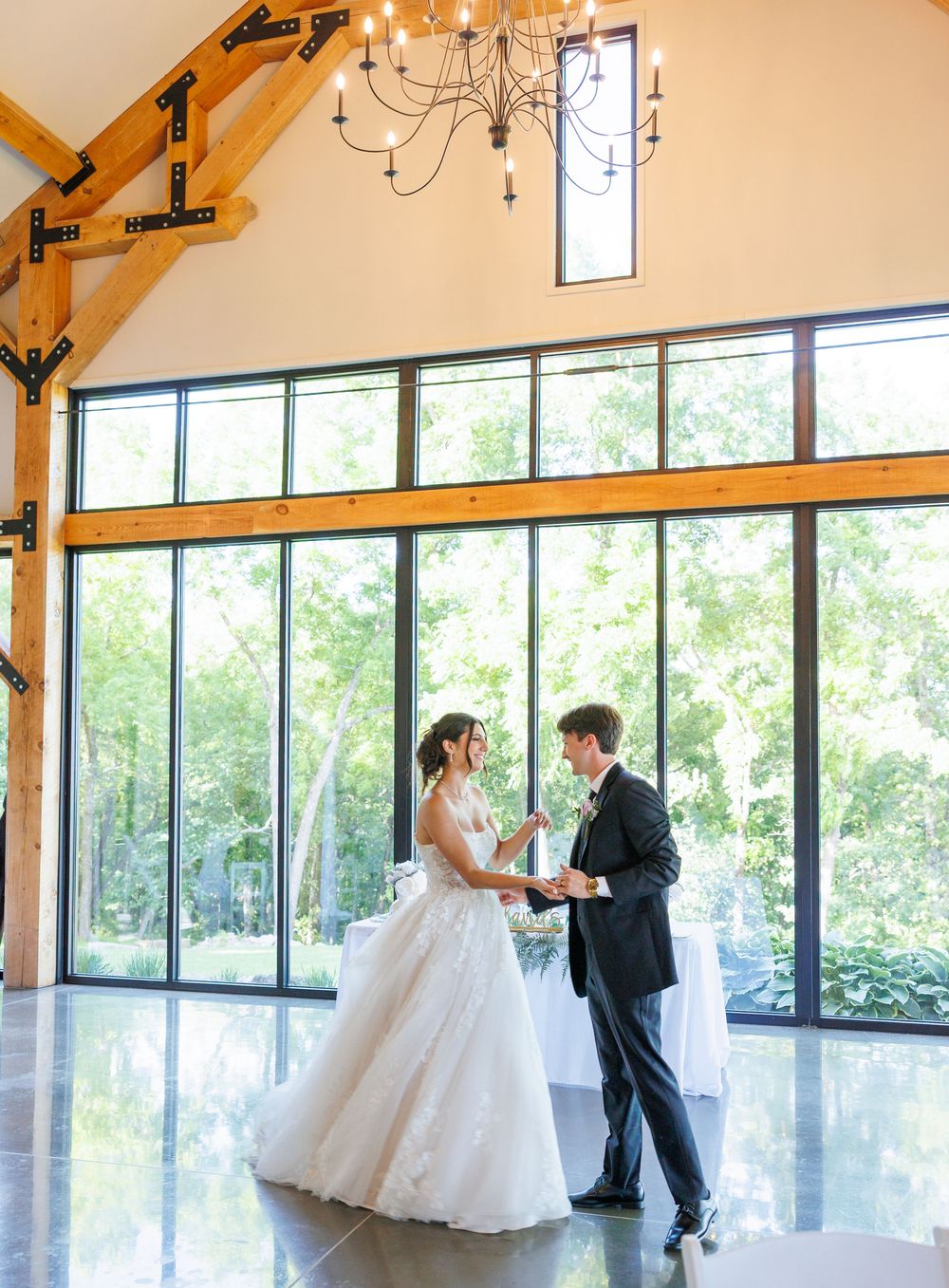 Couple shares first dance in elegant venue with floor-to-ceiling windows, wooden beams and chandelier overhead.