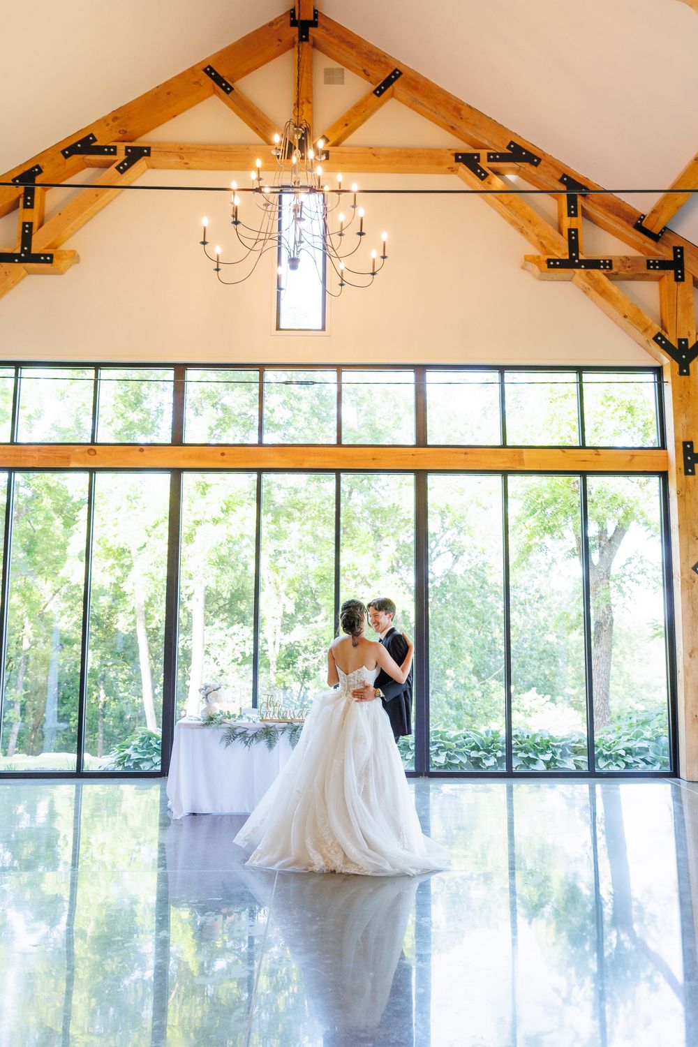 A light-filled wedding venue with exposed wooden beams and large glass windows shows dancing couples in white gowns.
