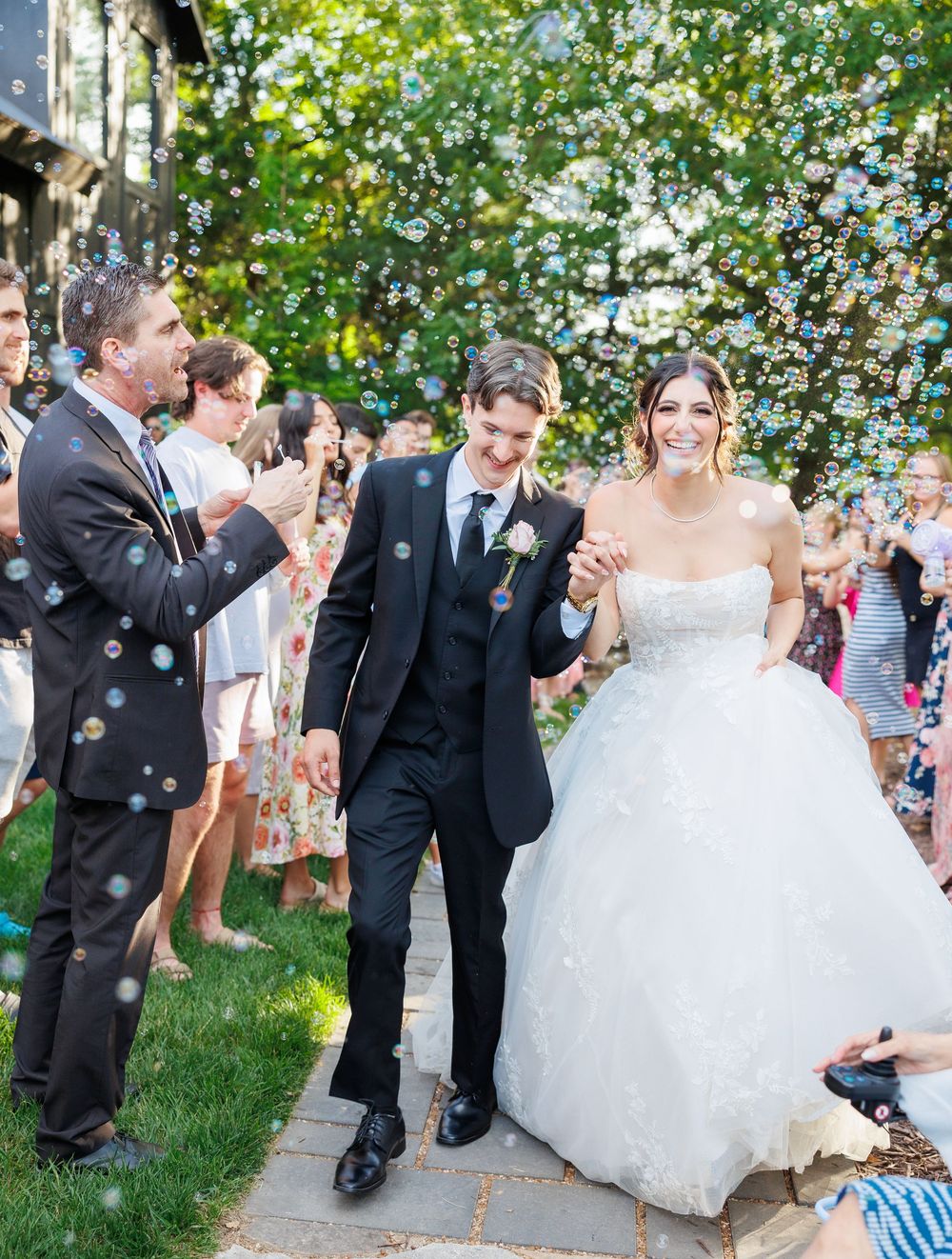 Wedding celebration with bubbles floating through the air as newlyweds walk down garden path surrounded by guests.