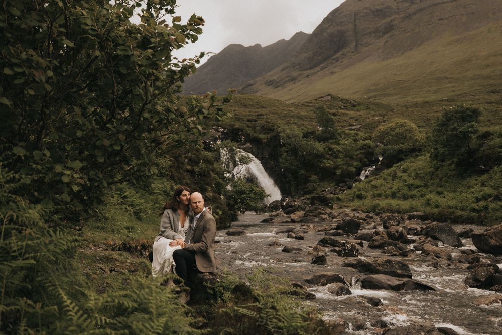 A couple embraces against a scenic backdrop of rugged Scottish mountains and flowing streams in Glen Coe.