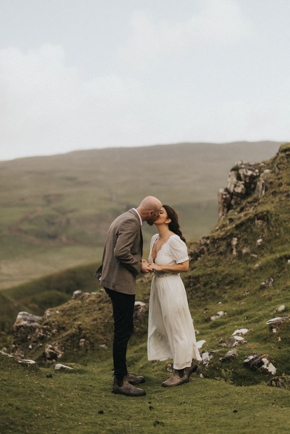 A couple shares an intimate moment on a rugged mountainside overlooking misty Scottish highlands.