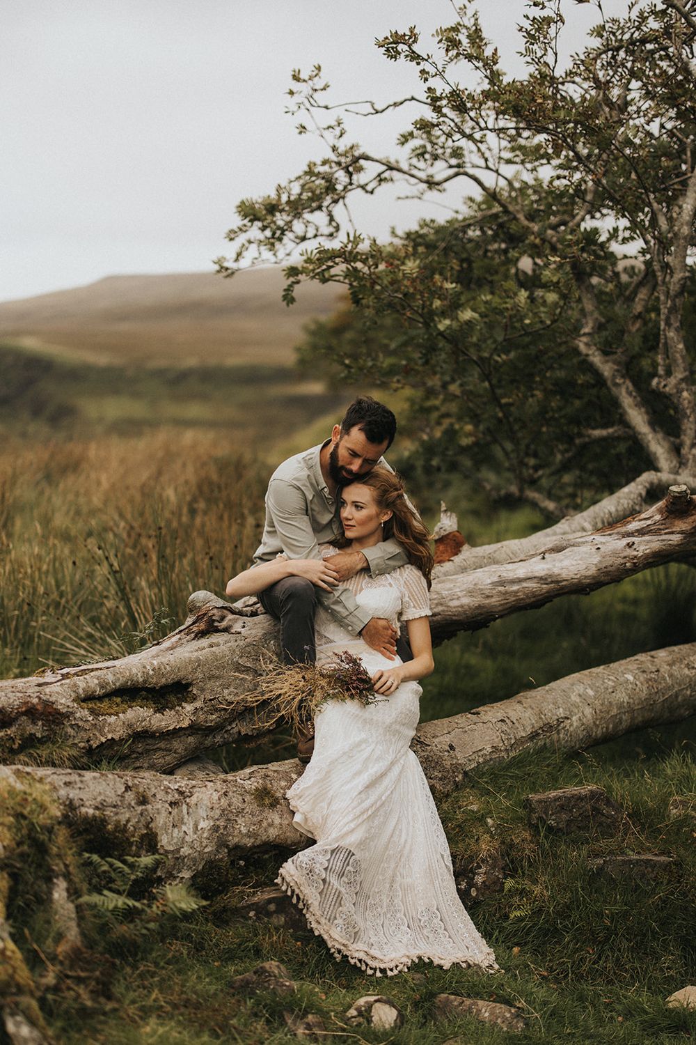 A romantic couple embraces on a fallen tree trunk in a scenic mountain landscape during golden hour.