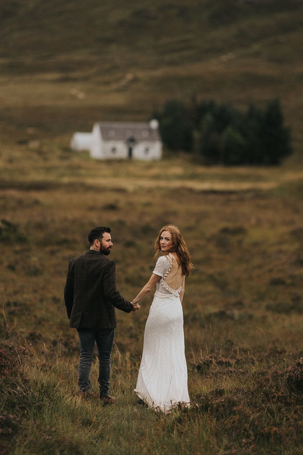 A couple in formal wedding attire walks through misty Scottish highlands with a white cottage in the distance.