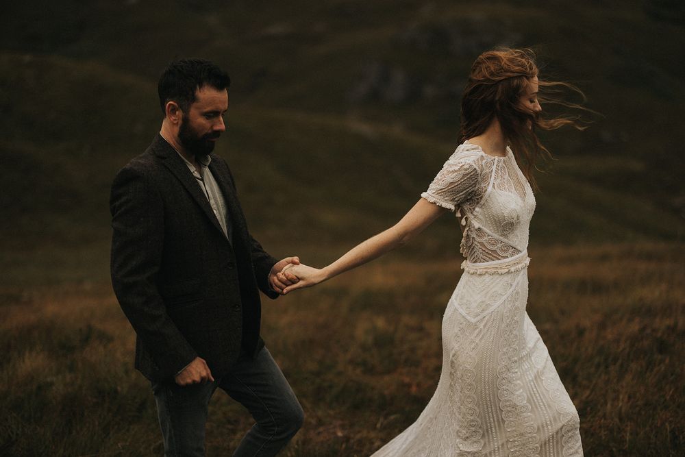 A couple in wedding attire walk through a misty field at sunset, with the bride's dress and hair flowing in the wind.