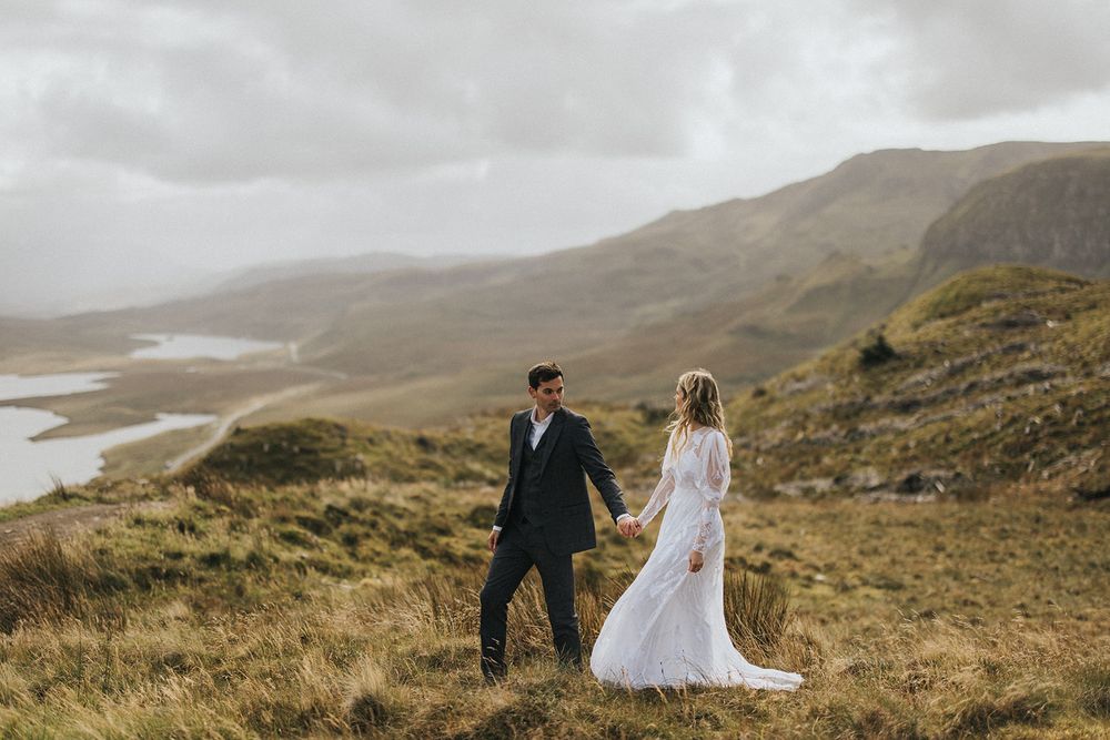 A couple in wedding attire shares an intimate moment on a misty Scottish hillside overlooking distant lakes and mountains.