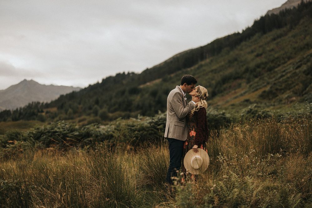A couple embraces on a misty mountainside with rolling green hills and cloudy skies in the background.