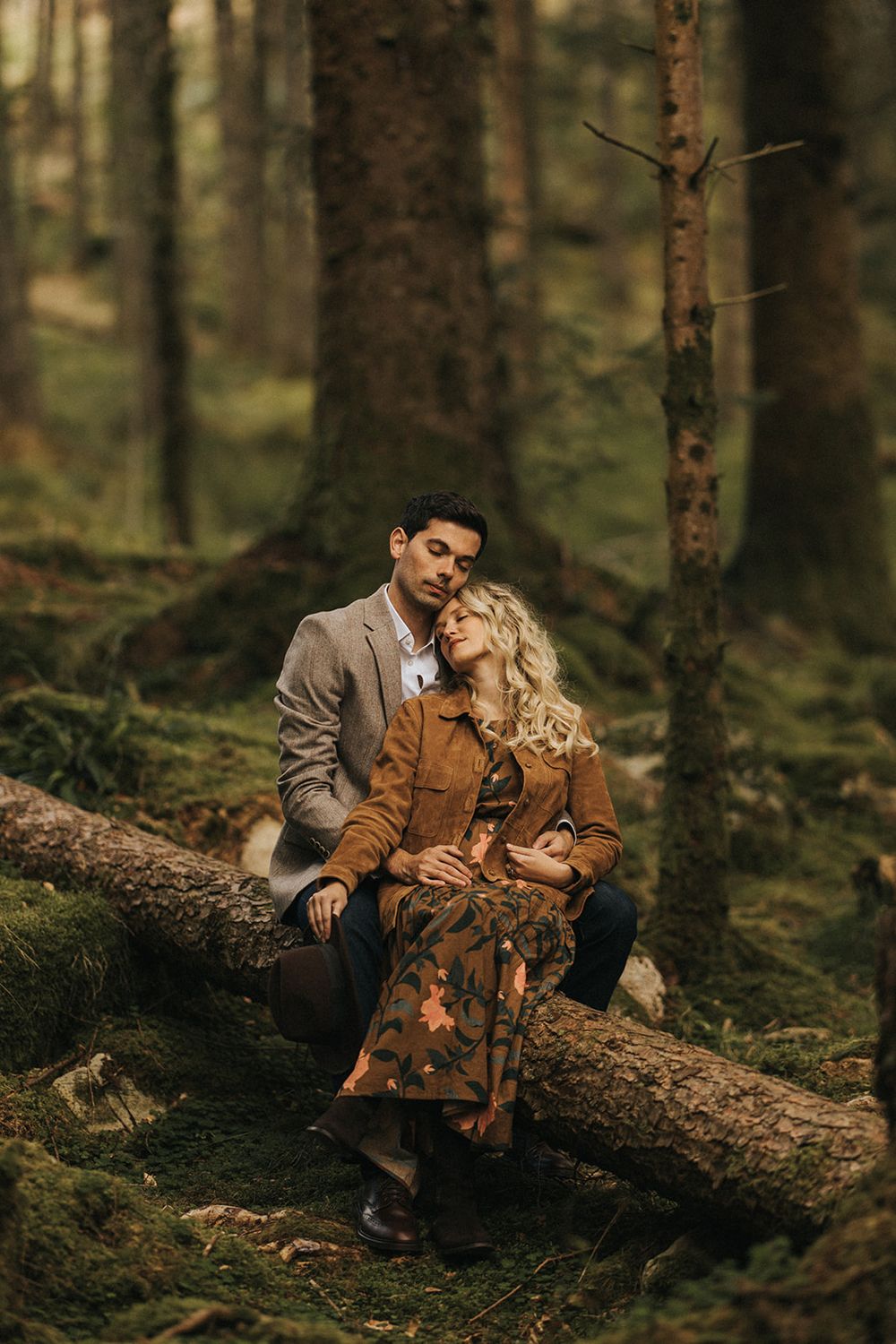 A couple in autumn attire sits intimately on a fallen log in a moody forest setting.