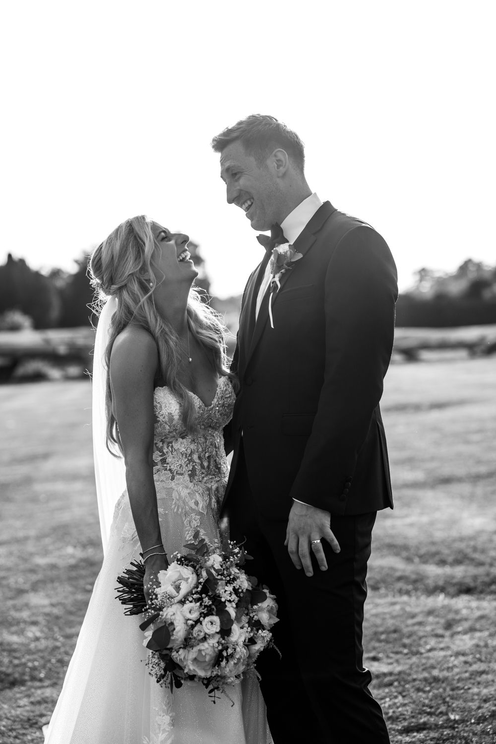 A black and white portrait of a joyful bride and groom sharing a candid laughing moment outdoors with wedding bouquet.