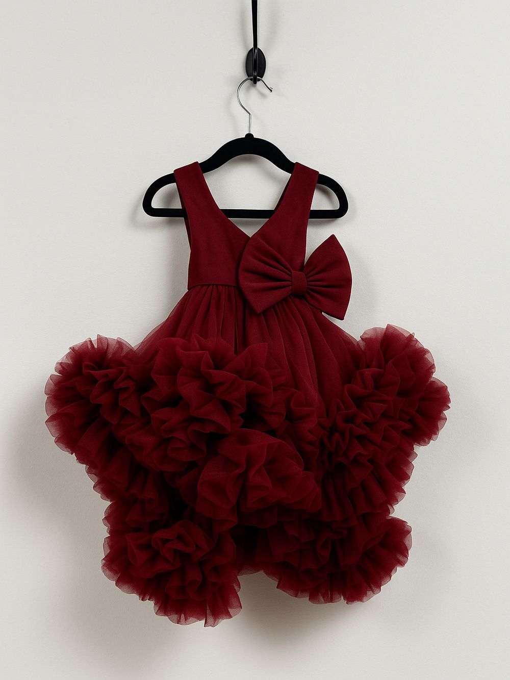 A burgundy red tulle dress with a large bow and dramatic ruffled skirt hangs on a black hanger against a white wall.