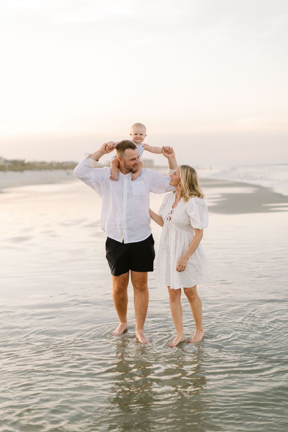 A family plays together in the shallow ocean water at sunset, wearing white and light colored clothing on the beach.