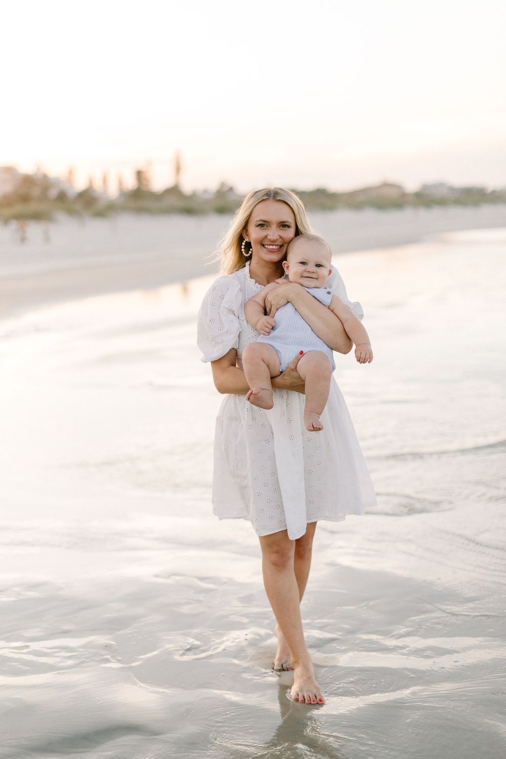 A parent and baby wearing white outfits walk along the beach at sunset with waves gently lapping at their feet.