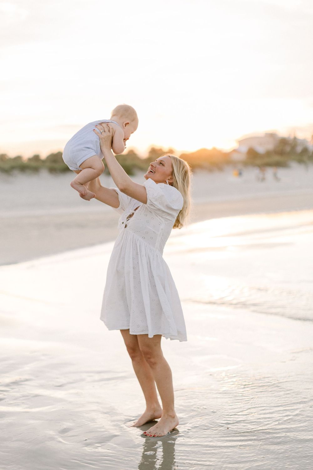 A person in a white dress walks along a sun-drenched beach shoreline during golden hour.