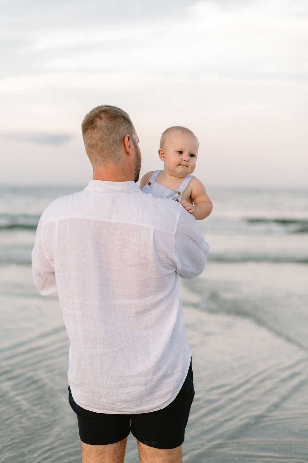 A figure in a white linen shirt holds a small baby while standing on a beach at sunset.