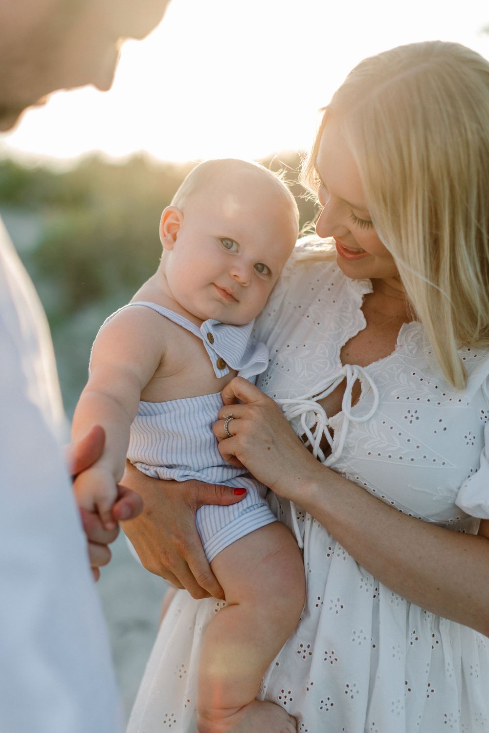 A small baby in striped clothing is held lovingly against a white eyelet dress on the beach.
