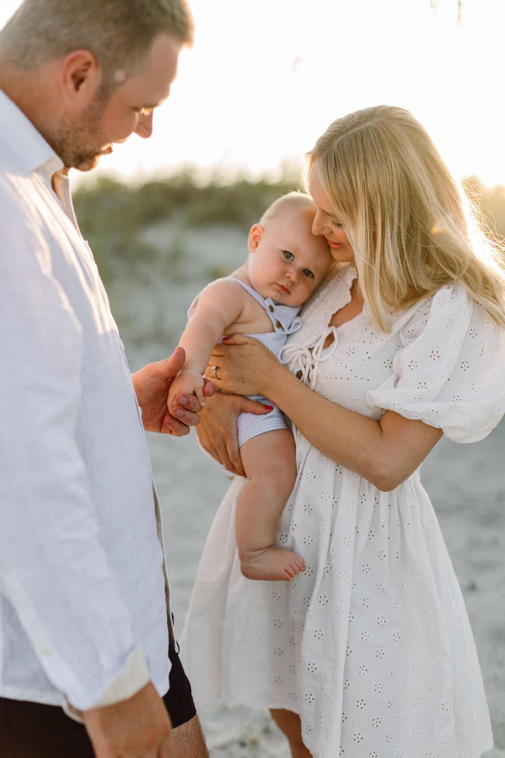 A couple in white clothing shares a tender moment with their baby on a sunny beach.