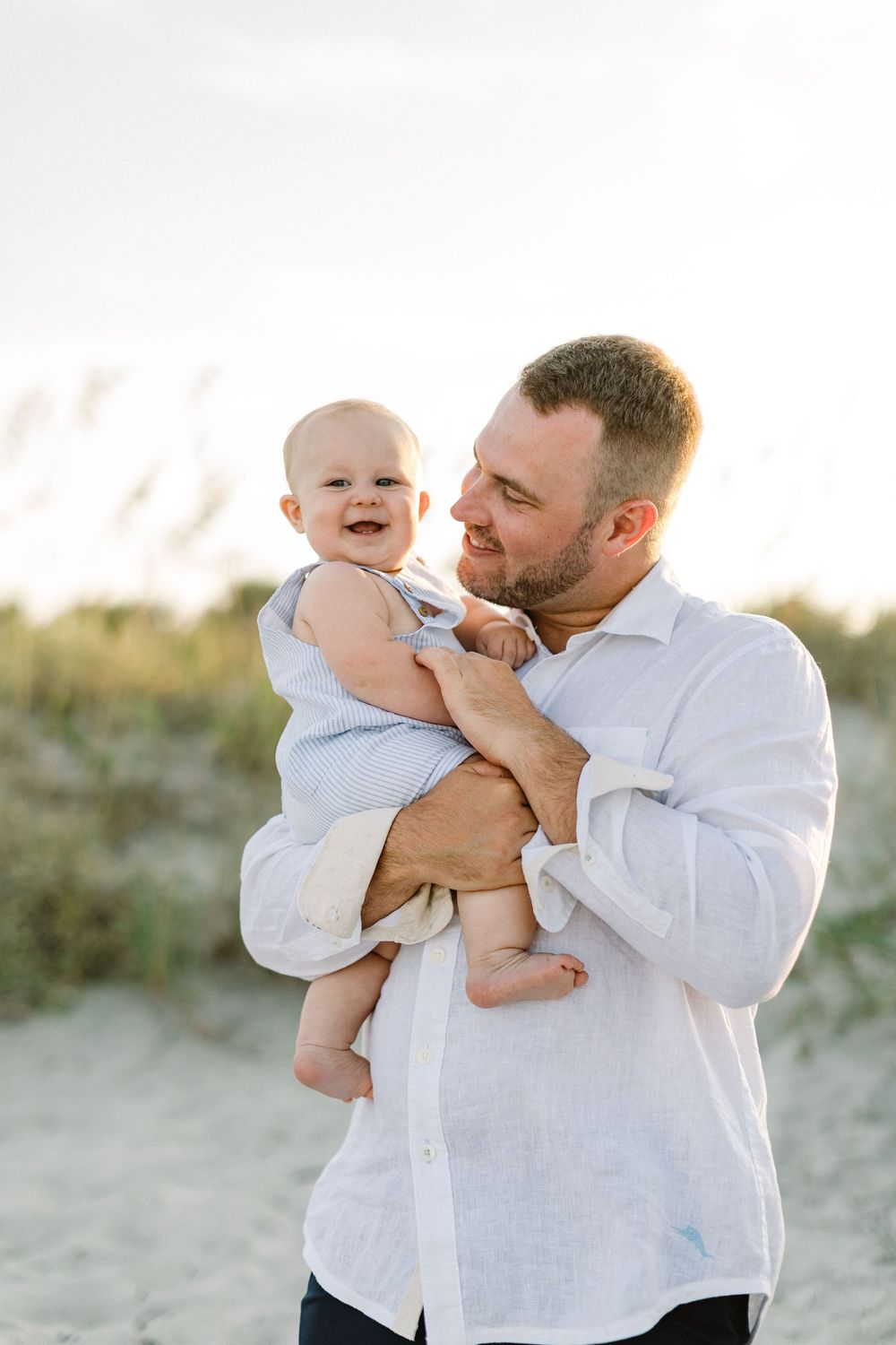 A heartwarming moment between an adult in a white shirt and a smiling baby at the beach during sunset.