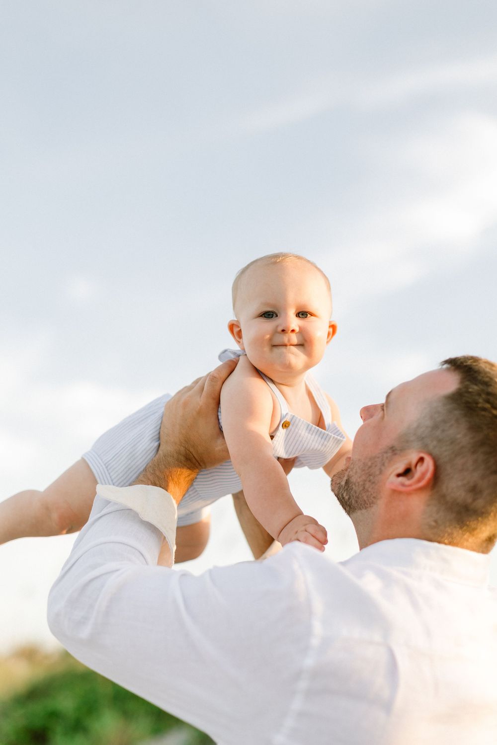 A baby in a white onesie smiles with joy while being lifted up against a bright blue sky.