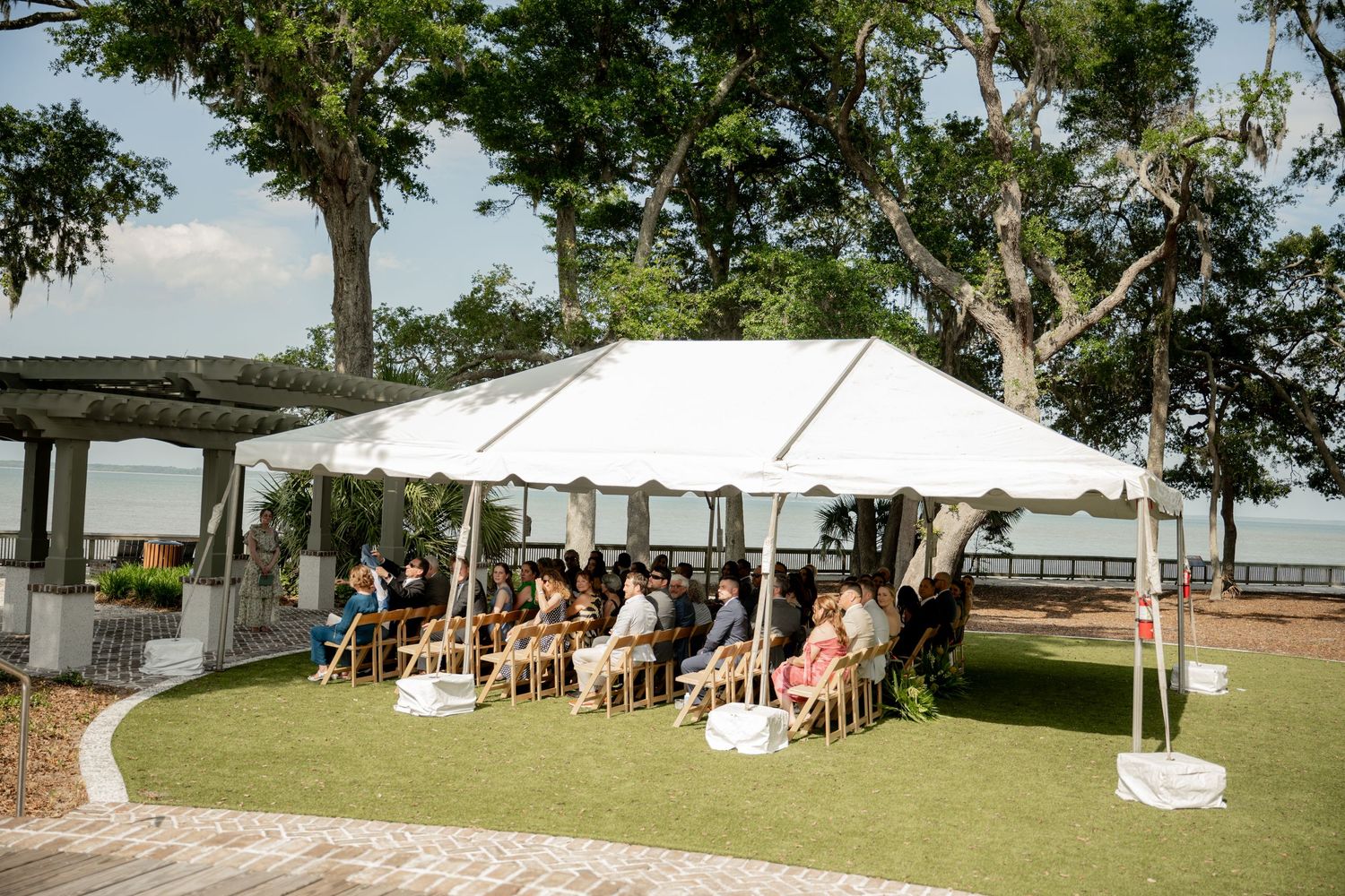 Outdoor wedding ceremony setup with white tent and chairs on a grassy lawn overlooking water and trees.